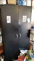 Front view of a tall black metal storage cabinet with two doors and two locking handles. Minor signs of use visible.