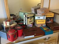 Photo shows an assortment of metal tins in various shapes and sizes along with wooden boxes including a wooden COHIBA box. Tins are branded and decorative.