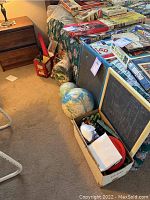 Overview of lot showing games stacked on bed, cardboard box with assorted toys including Rubik’s Cube, toy tank, chalkboard and globes.