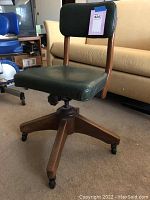 Full view of the vintage office chair showing wooden frame, green vinyl seat and back, and caster wheels.