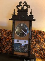 Full view of entire clock on patterned orange and black couch background showing wooden body, black clock face, day/date panels, lower glass section with pendulum and atomic design.