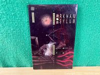 Front cover of the Batman: Arkham Asylum deluxe hardcover graphic novel showing title, writer, and artist names with a dark surreal art design.