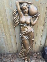 Full body view of bronze-tone nude female garden relief sculpture with vase on shoulder against wooden fence.