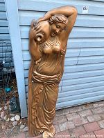 Full view of vintage nude female relief garden sculpture with bronze/golden finish standing against siding wall.