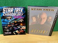 Photo showing two vintage Star Trek coffee table books standing upright on a green surface: 'Star Trek Chronology' with a blue cosmic cover and 'Star Trek Action!' with gray cover featuring Captain Picard and other characters.
