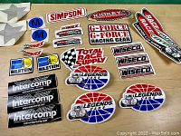 Overview of multiple vintage stock car racing stickers and patches arranged on table including brands Simpson, Kirkey, Wiseco, Intercomp, Bilstein, G-Force, Legends Cars, Total Race Supply and Race Fuel.