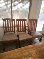Three wood dining chairs without armrests with vertical slat backs and beige upholstered seats arranged in a row in front of a window with blinds