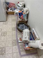 Overview of assorted cleaning supplies arranged on floor with baskets, including paper towels, replacement Swiffer heads, and small items.
