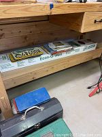 Long wooden shelf with box of 48 inch Philips Econ-O-Watt cool white fluorescent bulbs on top alongside assorted Oregon and Pennsylvania license plates lying flat. Various sandpaper sheets are visible in between license plates.