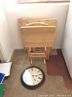 Four wooden folding trays stacked vertically with front handle visible and part of the folding rack below on carpet, clock face placed in front.
