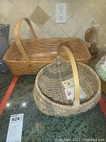 Two baskets on a countertop: a rectangular Longaberger basket with wooden adjustable handles and a handmade round Red Bird Mission basket with a woven fixed handle and paper tags.