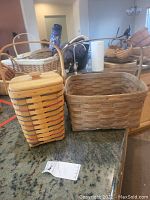 Two Longaberger baskets: one tall with lid and adjustable handle, one square with fixed handle on countertop.