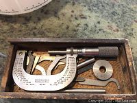 Photo of micrometer and attached measurement parts laid inside wooden box showing micrometer barrel, adjustment knob, several calibrating attachments, 1.000 calibration ring, and spanner wrench, all metal, wooden box interior worn.