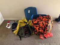 Overview of lot showing yellow rain jacket, orange camo clothing, gloves, hats, and blue storage bin.
