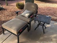 View of outdoor chair with metal frame, tan seat and back cushions, patterned throw pillow, matching ottoman with cushion, and decorative metal side table on concrete patio.