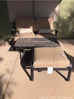 Outdoor loveseat, metal frame with beige cushions and two decorative throw pillows, matching ornate design metal table and ottoman with cushion shown from front angle.