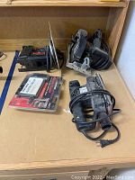 Four power tools on wooden surface: two jigsaws, circular saw, finishing sander, and electric stapler in packaging