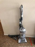 Full view of the silver Shark upright vacuum cleaner with attached hose and multiple hose attachments beside it on the floor in a tiled room.