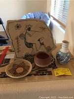 Flat slate plate with southwestern painted figure, two pottery pots including one signed pot with COA, and woven wicker basket arranged on beige cloth.