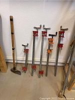 Seven woodworking steel clamps of various sizes arranged leaning against a wall next to the wooden handled metal-head cutter.