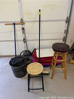 Photo shows two wooden stools, a large black plastic tote, and two brooms leaning against a garage door with a dust pan on floor.