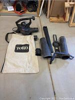 Photo showing TORO electric yard leaf blower with power cord, vacuum bag, various black plastic tubes and accessories laid out on floor.