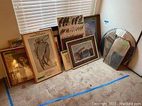 Overview photo showing assorted framed art prints and mirrors grouped on a carpeted floor against a wall with window blinds.