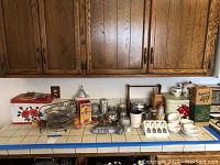 Full view of vintage kitchen items arranged on a tiled countertop including enamelware tin, metal trays, various glass and metal kitchen accessories, and ceramic items.