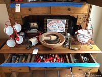 Wide view of wooden buffet surface showing enamelware rack, three-tier stand, wooden bowls, manual tools, tin sign and open drawers of utensils.