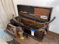 Wooden toolbox opened showing assorted vintage hand tools including wrench, hammer, brass blowtorch, clamp, brace drill and hand brush.