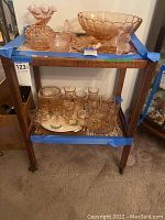 Front view of the wooden rolling 2-tier bar cart showing the top with various pinkish glass decorative pieces and the bottom with small glass cups and plates.