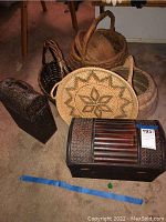 Wicker baskets and decorative wood storage boxes arranged on carpet, showing variety of sizes and styles including nested cylindrical cotton wrapped baskets, rectangular wood with inlay, and natural woven baskets.