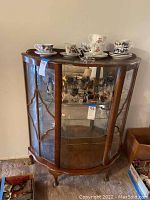 Front angled view showing curved top, side glass panels with wooden frames, decorative wood carving on front glass, and two interior glass shelves.