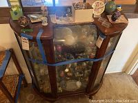 Full front view of curio cabinet showing wooden frame, glass panels, and blue tape securing it. Cabinet is filled with various collectibles (not included).