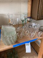 Overall view of the assorted glassware on a wooden shelf including plates, bowls, cups, and glasses