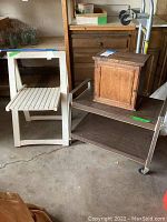 Photo showing metal rolling stand with two brown wood shelves on wheels, small white wooden folding chair, and medium brown wooden storage cabinet with door on top shelf of rolling stand.