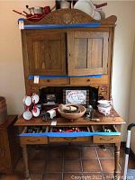 Front view of vintage wooden kitchen hutch with decorative crest, two cabinet doors, two small drawers, large lower drawer, and contents on workspace.