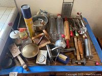 Top-down view of vintage kitchen tools laid out on a wooden surface, showing rolling pins, glass jars, hand-crank beaters, graters, and other assorted metal and wooden tools.