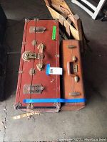 Photo showing two vintage suitcases, one large reddish-brown with metal locks and one smaller brown with combination lock, alongside a canvas backpack folded on the floor.