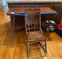 Photo shows two vintage wooden square tables with turned legs and a vintage wooden rocking chair with cane seat and backrest in front. Items placed on hardwood floor.