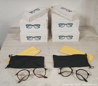 24 white retail boxes stacked with black storage pouches and yellow cleaning cloths, two glasses pairs displayed in front (tortoiseshell and black round frames)