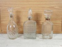 Front view of three various-shaped antique glass decanters with their corresponding glass stoppers on a wooden background.