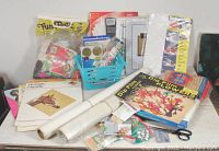 Wide view of the crafting supplies lot including multiple books, white paper rolls, and various crafting materials.