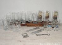 Photo showing multiple clear beer glasses with logos from different breweries, a wooden Turtle Island sampler tray, and metal bottle openers arranged on a surface.