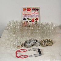 Lot of clear glass Mason jars in various sizes, multiple metal lids and rings, jar tongs with red grips, and a canning guidebook standing behind the jars.