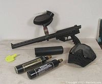 Black Stingray paintball marker with attached hopper, two CO2 tanks, black protective mask, yellow barrel plug, and black paintball canister on a light wood surface.