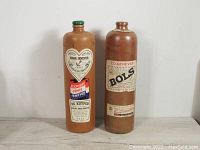 Two vintage stoneware gin bottles standing side by side on wooden surface against white background, showing labels and overall condition.