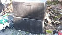 Two brown plastic outdoor storage lockers stacked outdoors on gravel ground near bricks and wood.