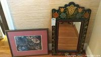 First image shows framed floral artwork and tole painted framed mirror side by side on wooden floor.