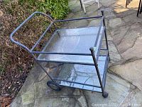 Full view of the patio bar cart showing metal frame, top glass shelf, handle, and rear wheels. Rust and peeling paint visible on metal parts.